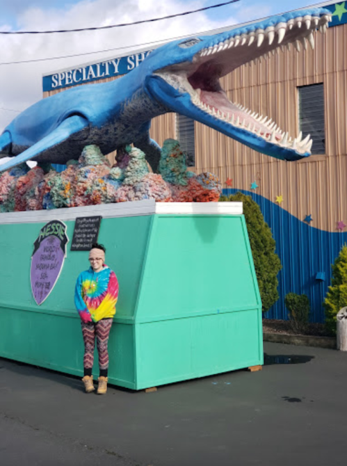 A person in a tie-dye shirt stands next to a large blue whale statue with an open mouth, surrounded by colorful coral.