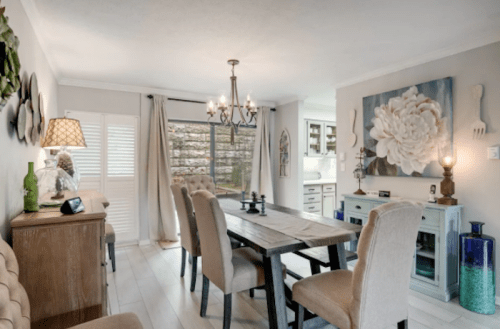 A modern dining room with a wooden table, beige chairs, a chandelier, and decorative wall art. Natural light from a window.