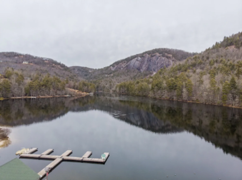 A serene lake surrounded by mountains and trees, with a dock extending into the calm water under a cloudy sky.