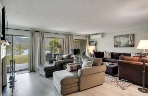 Cozy living room with large windows, comfortable seating, and a view of a lake and greenery outside.