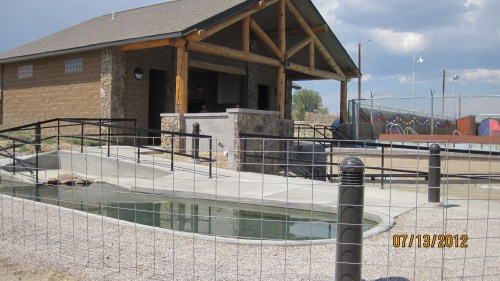 A building with a stone facade near a fenced area, featuring a pool and playground in the background under a cloudy sky.