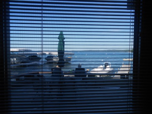 View of a serene lake scene through window blinds, featuring boats and a green umbrella on the dock.