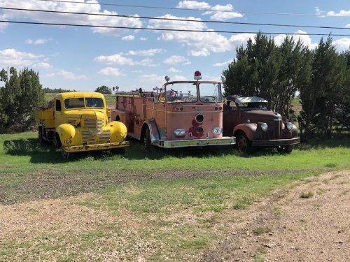 Three vintage vehicles parked on grass: a yellow truck, a pink fire truck, and a brown truck, with blue skies above.