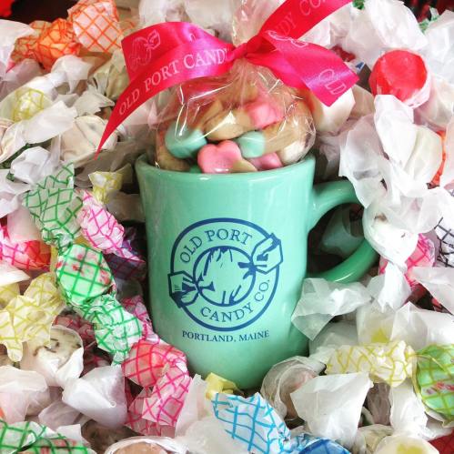 A green mug with "Old Port Candy Co." logo filled with colorful candies, surrounded by crinkled candy wrappers.