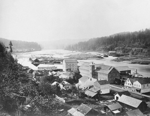 Historic black-and-white view of a river town with buildings and forested hills in the background.
