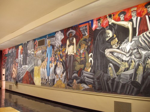 A vibrant mural depicting historical and social themes, featuring figures, skeletons, and scenes of struggle and resilience.