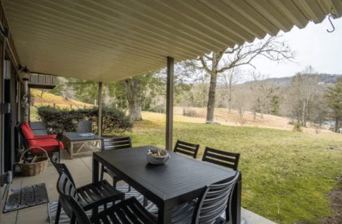 A covered patio with a dining table and chairs, overlooking a grassy area and trees in the background.