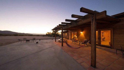 A rustic wooden porch with a hammock, overlooking a desert landscape at dusk. Warm lights illuminate the space.