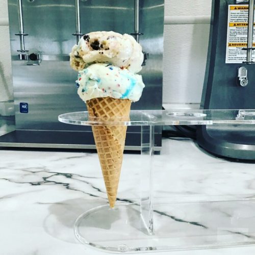 A colorful ice cream cone with two scoops, one cookie dough and one blue, displayed on a clear stand.