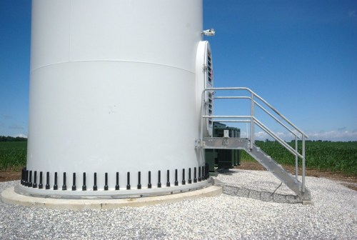 A wind turbine base with a metal staircase leading up, set against a clear blue sky and green fields.