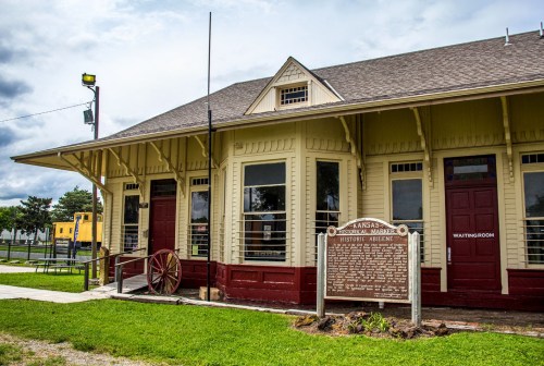 Historic train station with a sign, featuring a yellow exterior and red doors, surrounded by greenery and a cloudy sky.