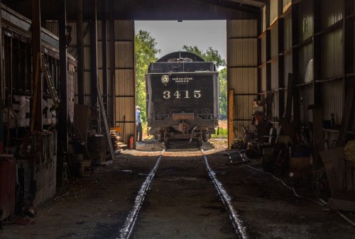 A vintage steam locomotive sits in a dimly lit shed, with tracks leading out into bright daylight.