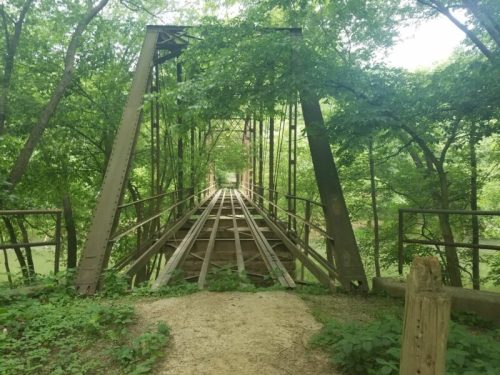 A weathered metal bridge surrounded by lush green trees, leading over a calm river.
