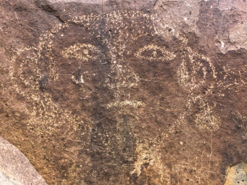 Petroglyph of a human face etched into a rock surface, featuring distinct eyes and ear-like shapes.