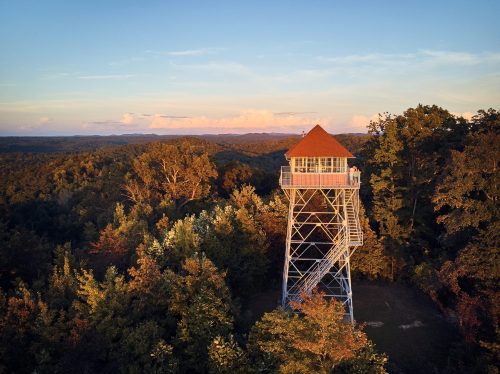 A tall fire tower stands amidst lush trees, overlooking a scenic landscape at sunset.