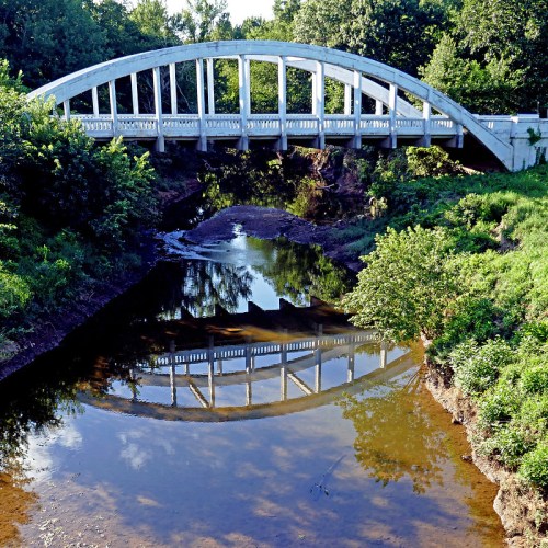 A white arch bridge spans a calm river, reflecting its shape in the water, surrounded by lush greenery.