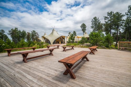 A wooden deck with benches overlooks a tent-like structure surrounded by trees under a cloudy sky.