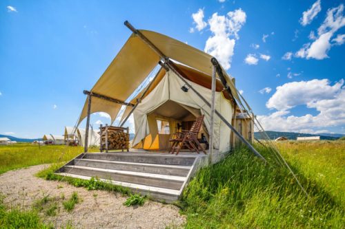 A spacious glamping tent with a wooden deck, surrounded by green grass and blue skies with fluffy clouds.