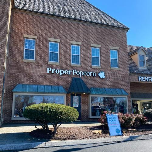 Exterior of Proper Popcorn shop with a sign indicating "We're Open" and a sunny blue sky in the background.