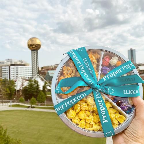 A hand holds a tin of colorful popcorn with a city skyline and a golden sphere in the background.