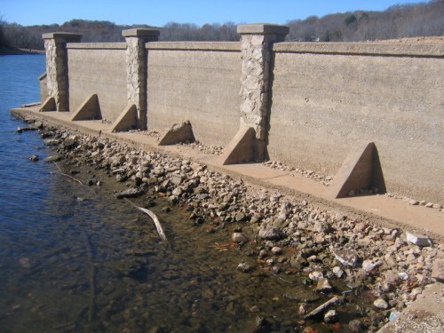 A stone wall along a water's edge, with support pillars and a rocky shoreline visible.