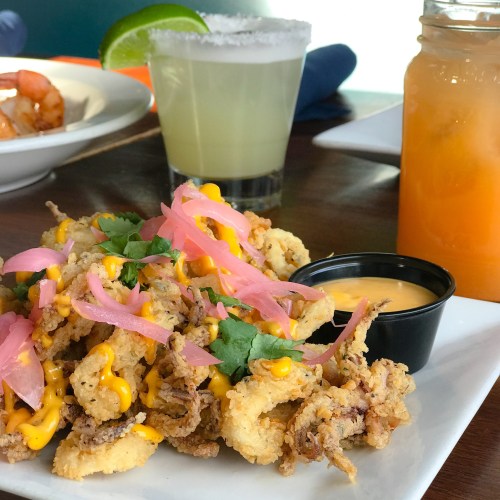 A plate of crispy calamari topped with cheese, pickled onions, and cilantro, served with a dipping sauce and drinks in the background.