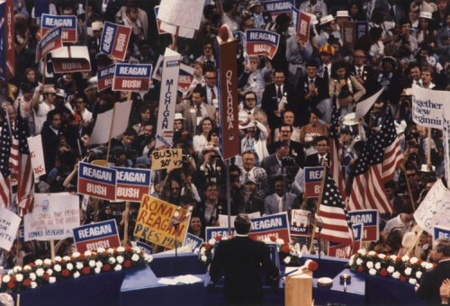 A crowd at a political rally holds signs for Reagan and Bush, with American flags and a speaker at the podium.