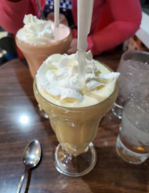 Two tall glasses of milkshakes topped with whipped cream, served on a wooden table with a spoon and a glass of water.