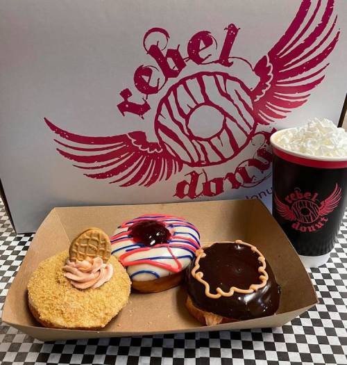 Three colorful donuts on a tray with a coffee cup topped with whipped cream, set against a branded box.