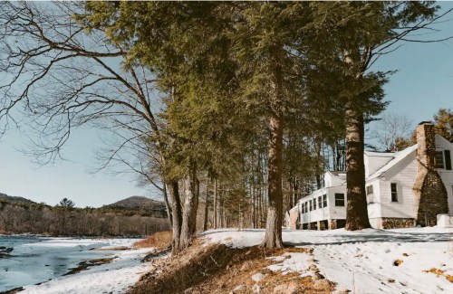 A serene winter landscape featuring a white house by a snowy riverbank, surrounded by tall trees and mountains in the background.