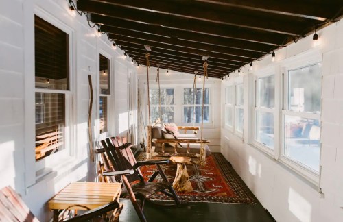 A bright sunroom with wooden beams, cozy seating, and a patterned rug, featuring large windows and warm lighting.