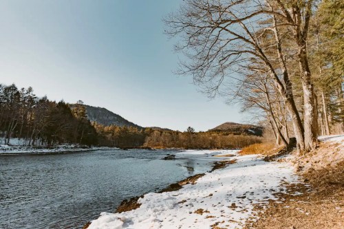 A serene river scene with snow-covered banks, bare trees, and mountains in the background under a clear blue sky.