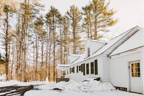 A white house surrounded by tall pine trees, covered in snow, with a clear sky in the background.