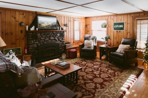 Cozy living room with wooden walls, stone fireplace, two armchairs, and a decorative rug. Natural light from windows.