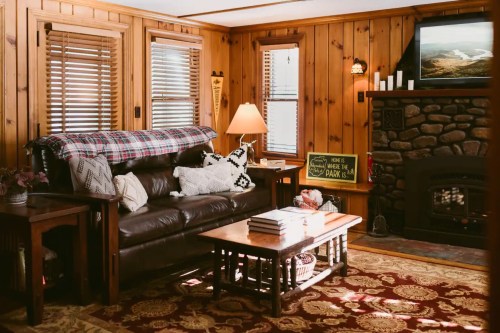 Cozy wooden living room with a leather sofa, coffee table, and stone fireplace, featuring warm lighting and rustic decor.