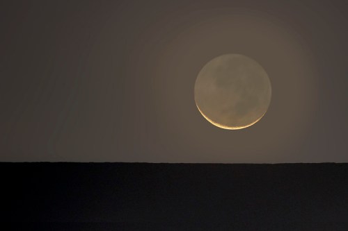 A crescent moon rises above a dark horizon, casting a soft glow against a twilight sky.