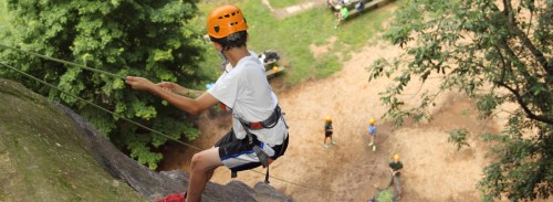 A young climber in an orange helmet rappels down a rock face, with others climbing in the background.