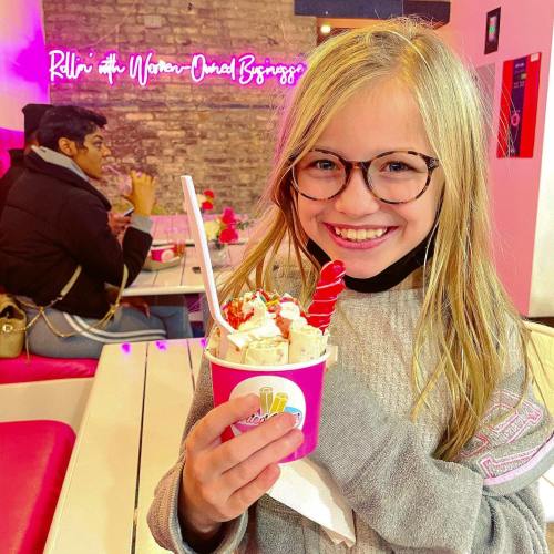 A smiling girl with glasses holds a colorful ice cream cup in a vibrant café setting.