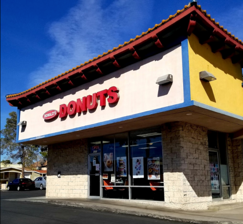A storefront of "Ronald's Donuts" featuring a bright sign and large windows, with a clear blue sky above.