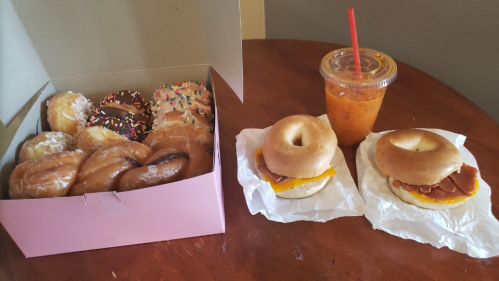 A box of assorted donuts next to two bagel sandwiches and an iced drink on a wooden table.