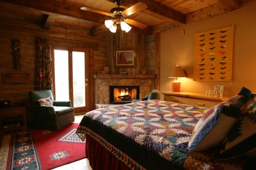 Cozy bedroom with a quilted bed, fireplace, and rustic decor, featuring wooden beams and colorful artwork.
