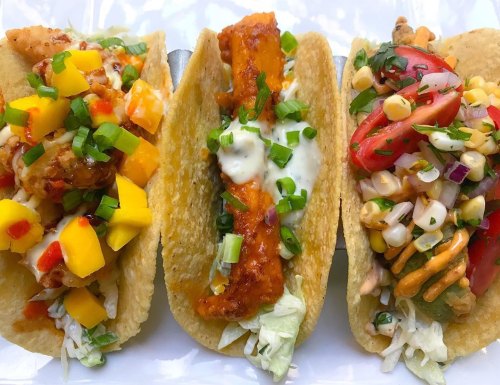 Three colorful tacos filled with various toppings: mango salsa, crispy fish, and corn salad, garnished with green onions.