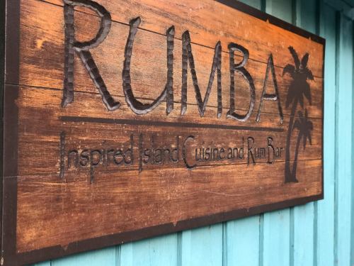 Sign for "Rumba" featuring carved letters, palm tree design, and the tagline "Inspired Island Cuisine and Rum Bar."