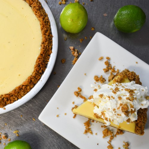 A slice of lime pie topped with whipped cream, next to a whole pie and fresh limes on a silver surface.