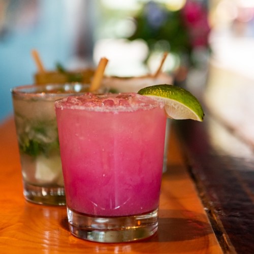 Three colorful cocktails on a wooden bar, featuring a pink drink with a lime wedge in the foreground.