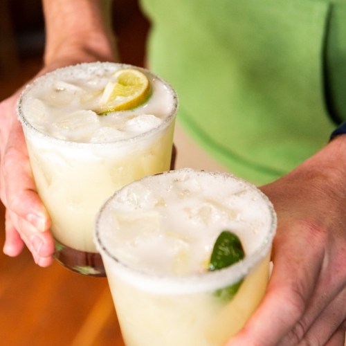 Two glasses of margaritas with lime slices, held by a person wearing a green shirt.