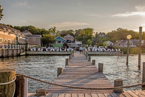 A wooden dock extends into calm water, leading to a waterfront restaurant surrounded by trees and houses.