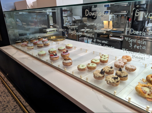 A display case filled with various colorful donuts and pastries, labeled with flavors and descriptions.