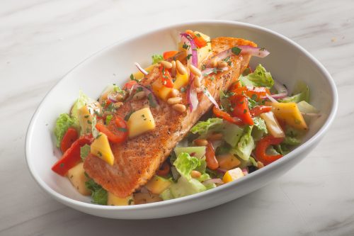 A bowl of salad topped with grilled salmon, mango, red peppers, and pine nuts on a bed of lettuce.