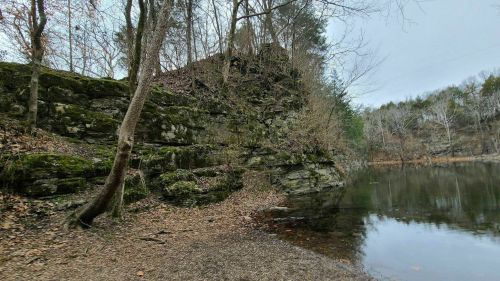 A serene lakeside scene with rocky cliffs, bare trees, and a calm water surface reflecting the surroundings.